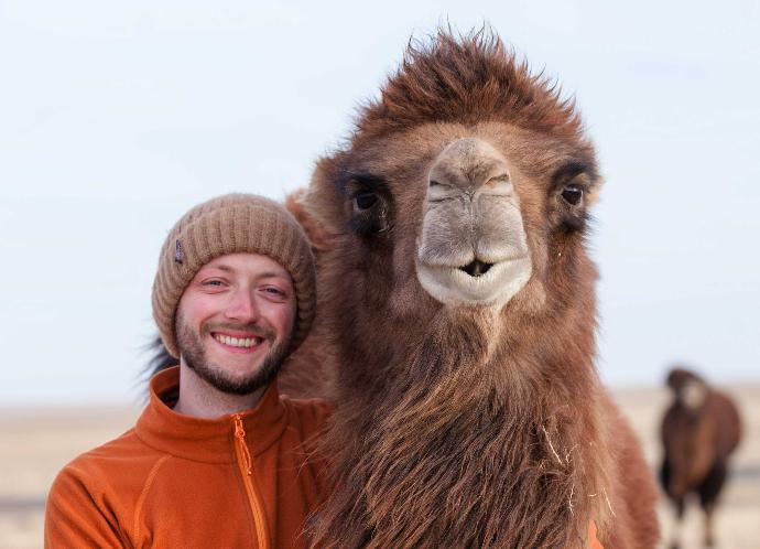Malte mit brauner Mütze lächelt neben Kamel in die Kamera