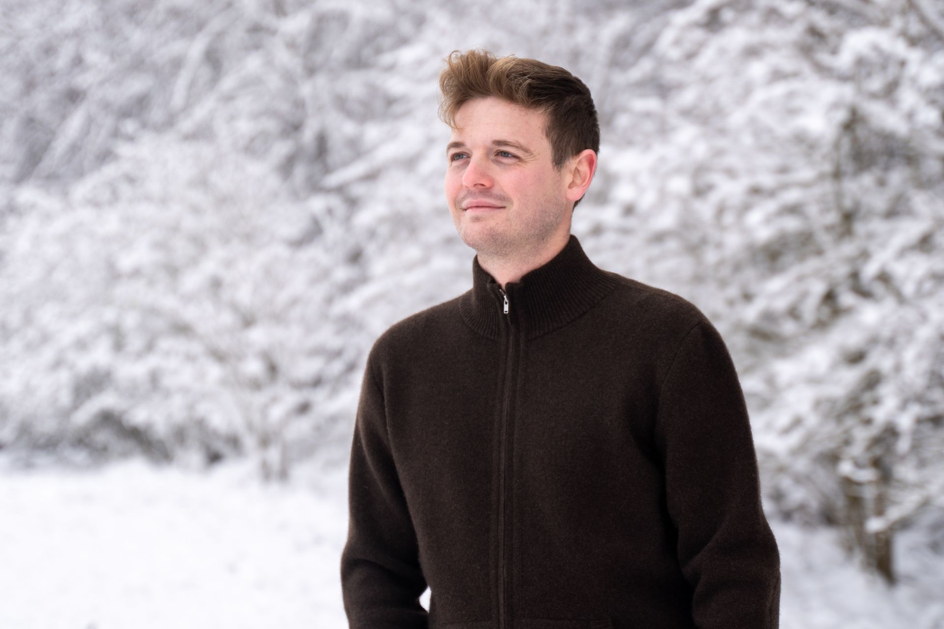 Young man in a dark cardigan stands in front of a snow-covered forest