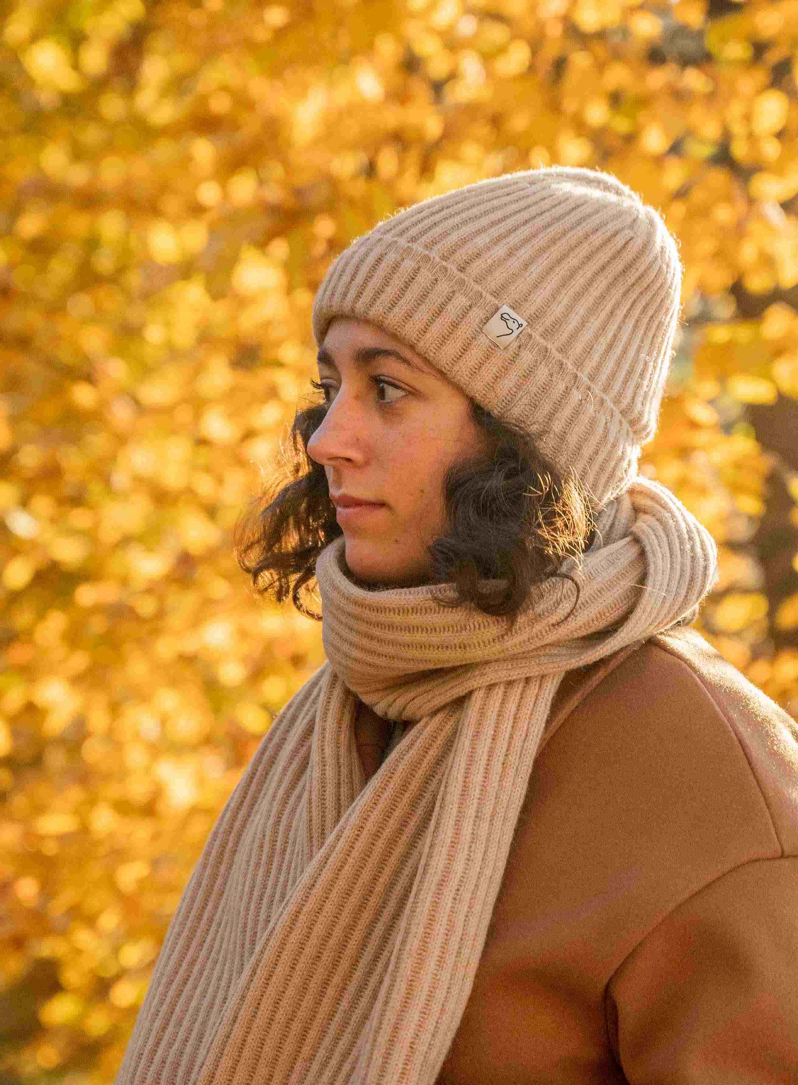 Frau mit beigem Schal und Mütze aus Kamelwolle vor einer orangenen Hecke