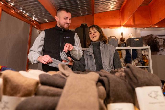 Zwei Personen in warmen Winterkleidern hinter einem Stand mit Wollprodukten, lächelnd im Gespräch.