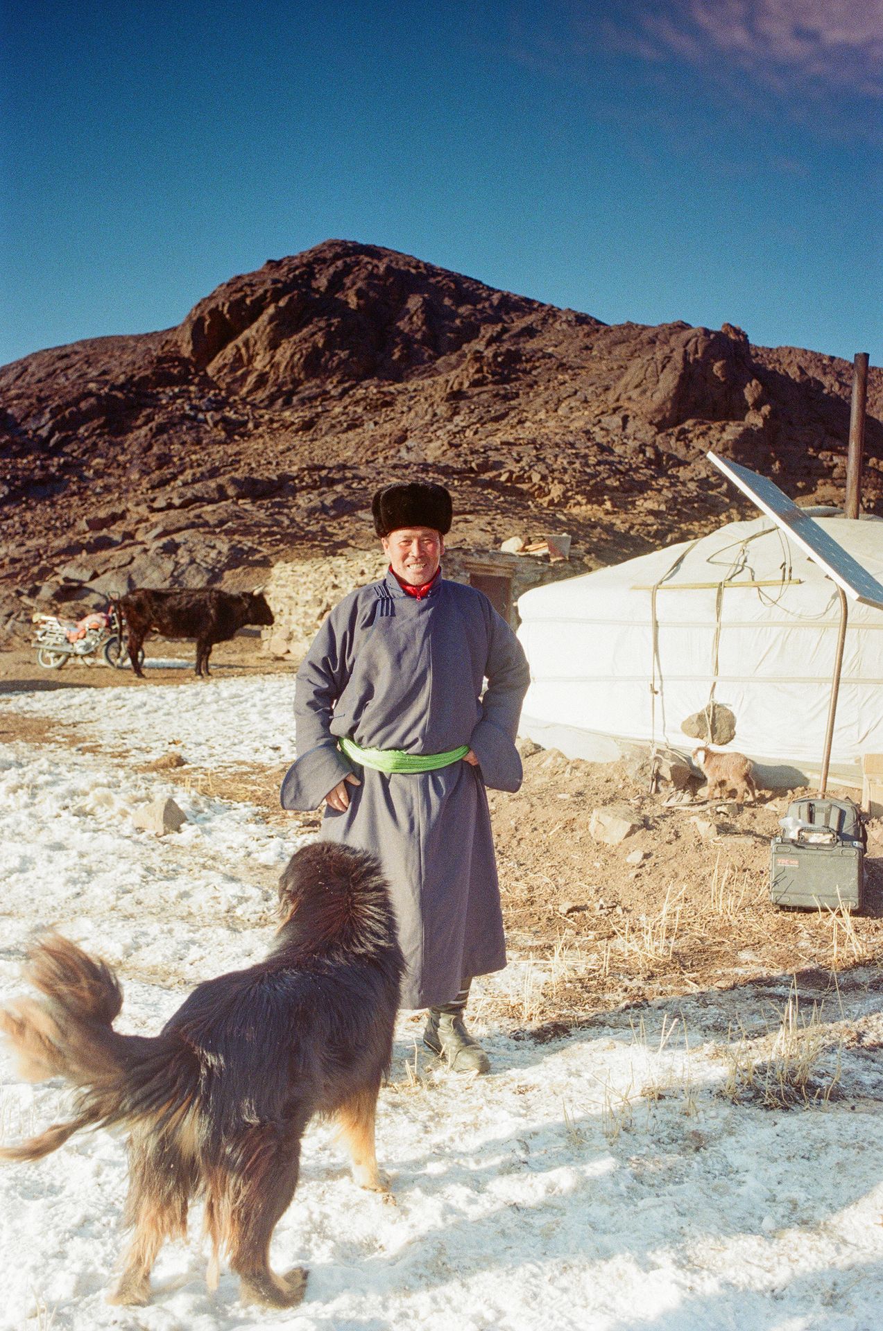 Mann in traditioneller Kleidung lacht in die Kamera, ein Hund steht vor ihm – Jurte und Berge im Hintergrund.