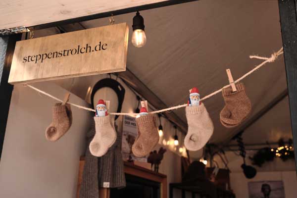 Top view of small, knitted wool socks in various colors hanging on a line with small clothes pegs under a steppenstrolch.de logo.