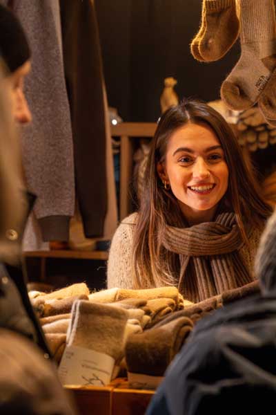A young woman with brown hair and a beige scarf smiles as she stands behind a stall selling knitted goods at a market.