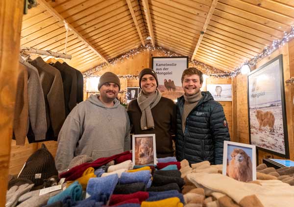 Three men are standing in a bright wooden hut in front of a display of socks, laughing.
