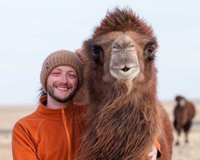 Ein junger Mann mit Mütze steht neben einem Kamel und beide schauen in die Kamera.